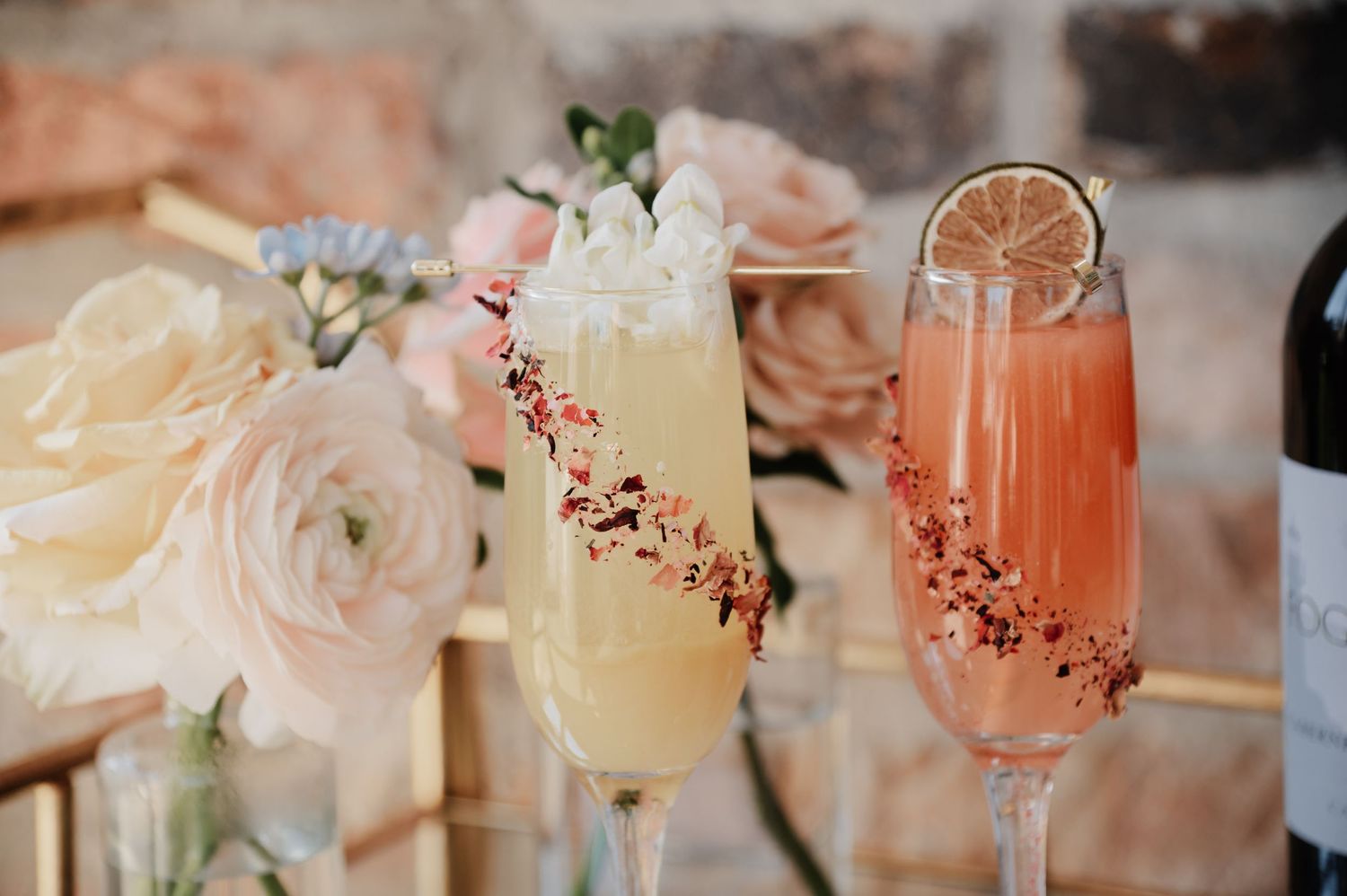 Two elegant champagne flutes with floral garnishes, one yellow and one pink mimosa, surrounded by soft pink roses.