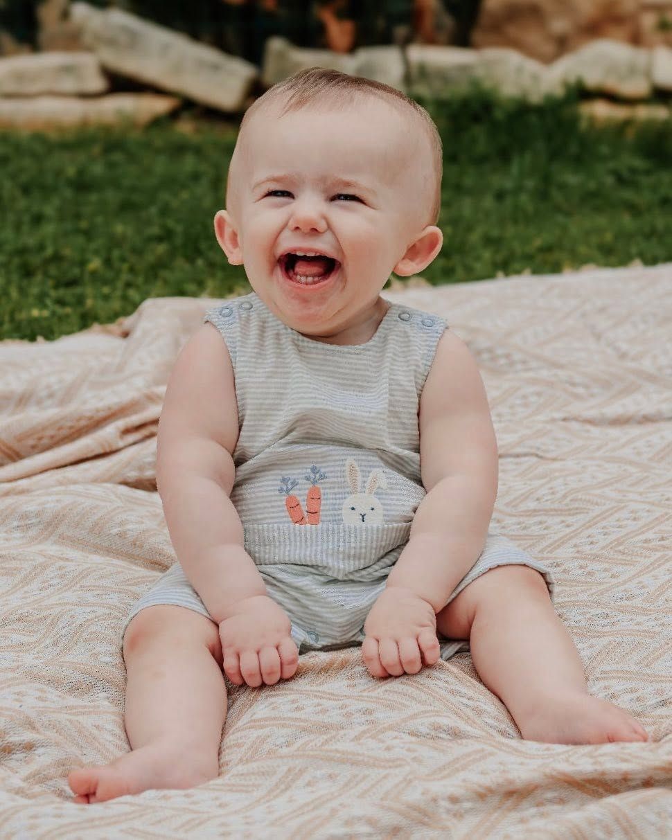 Happy baby in bunny romper sits on blanket outdoors, smiling joyfully with green grass background.