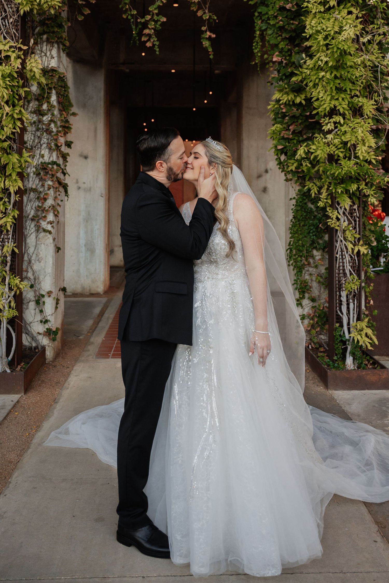 Bride and groom sharing first kiss in white ballgown and black tuxedo before ivy-covered venue entrance.