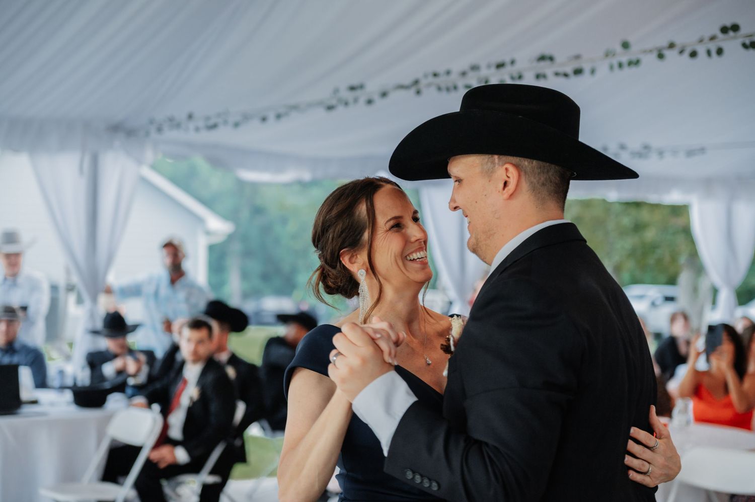 Groom in a black cowboy hat shares a joyful mother-son dance at an elegant tented wedding reception.