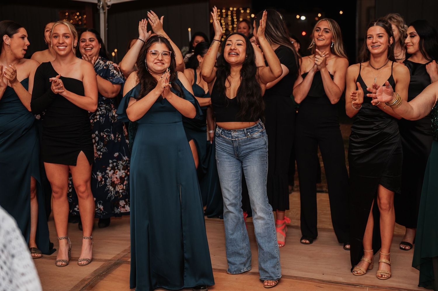 Group of women in elegant attire dancing and clapping joyfully at a party or wedding reception.