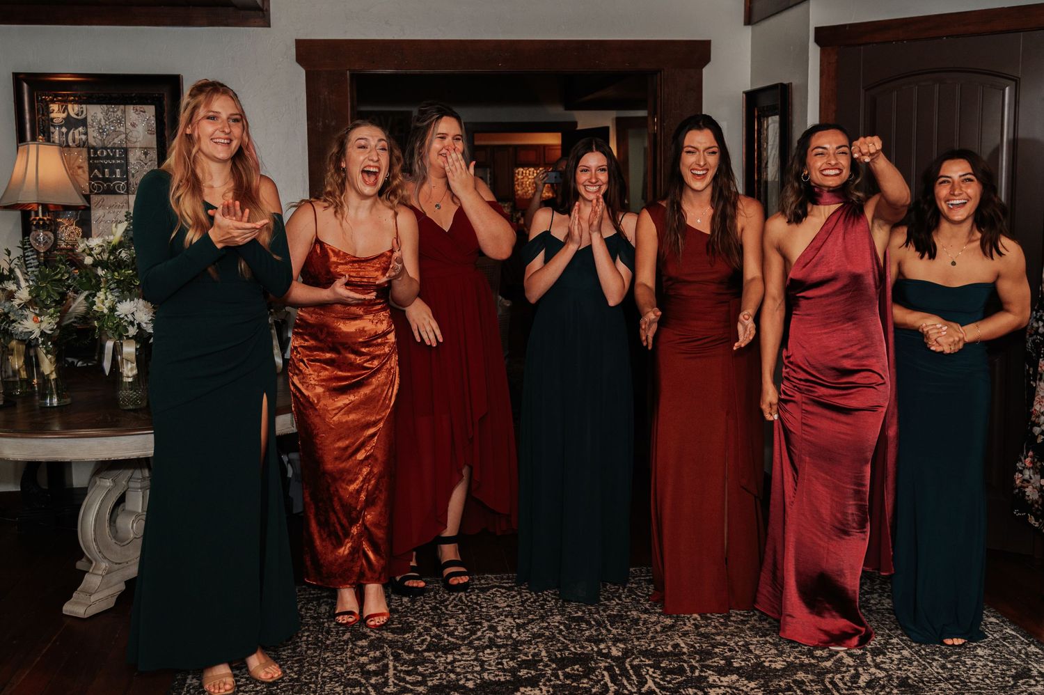 Group of bridesmaids in elegant satin gowns reacting with joy and excitement at a wedding celebration indoors.