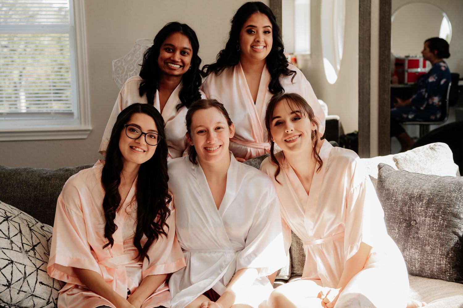 Five women in satin robes smiling together on a couch during bridal party getting ready preparations.