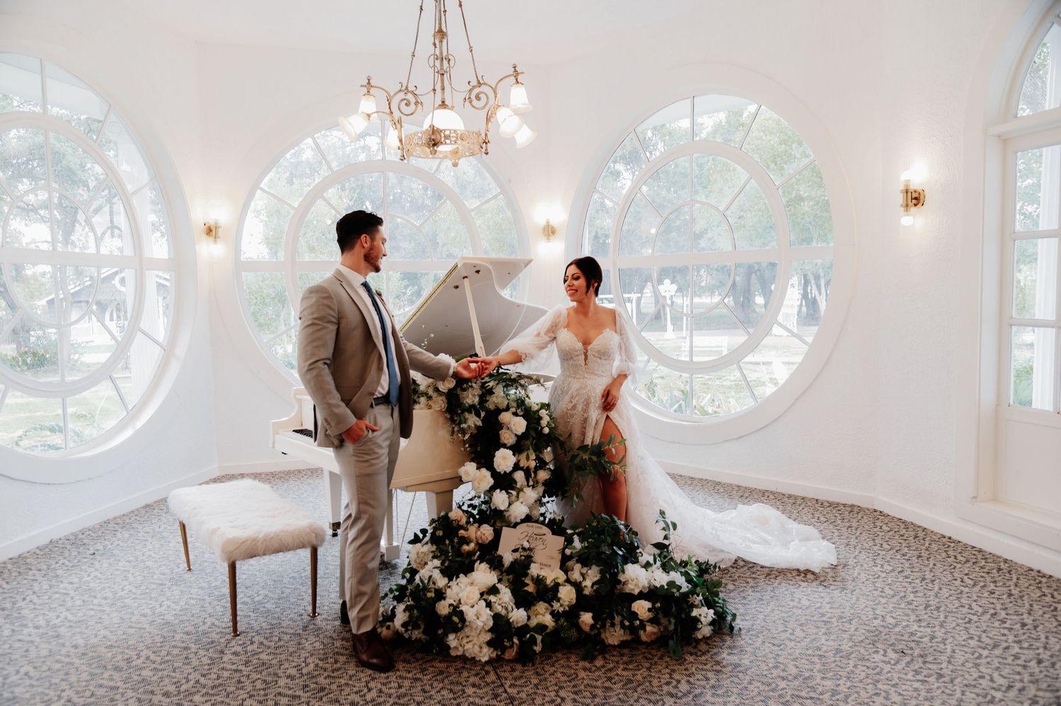 Bride and groom stand by a floral-adorned white grand piano in an elegant venue with arched windows.