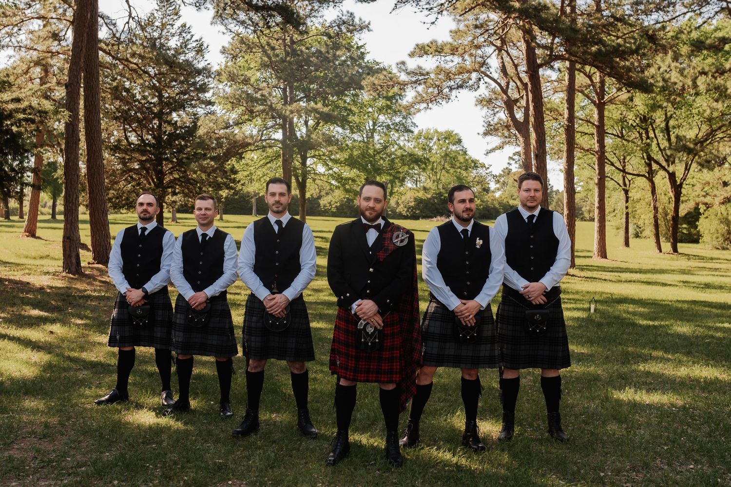 Six groomsmen in traditional Scottish kilts and black vests stand together outdoors on green grass surrounded by tall pine trees.