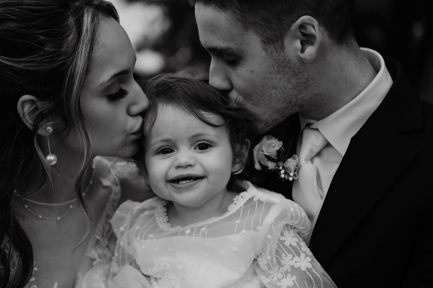 Black and white wedding photo of bride and groom kissing smiling baby girl in white dress.
