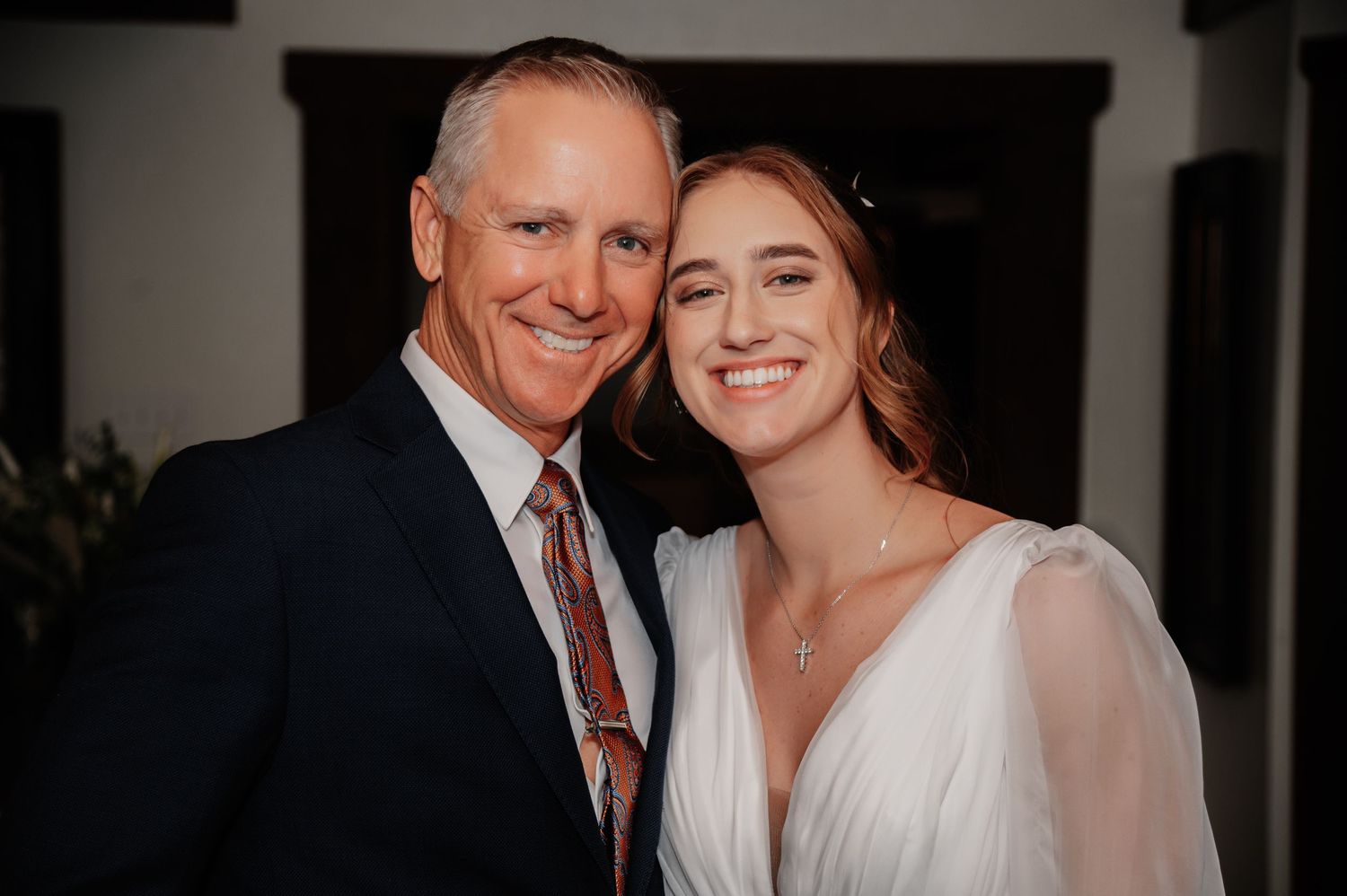A smiling bride in white dress poses with an older man in navy suit and floral tie at a wedding celebration.