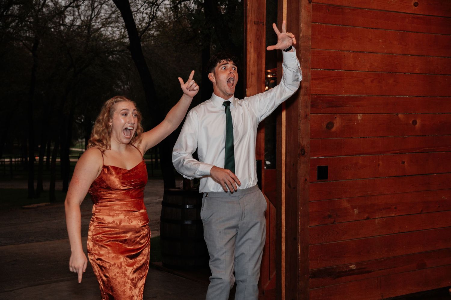 Excited couple in formal attire celebrating outside a rustic barn venue, woman in red dress and man in tie pointing up.