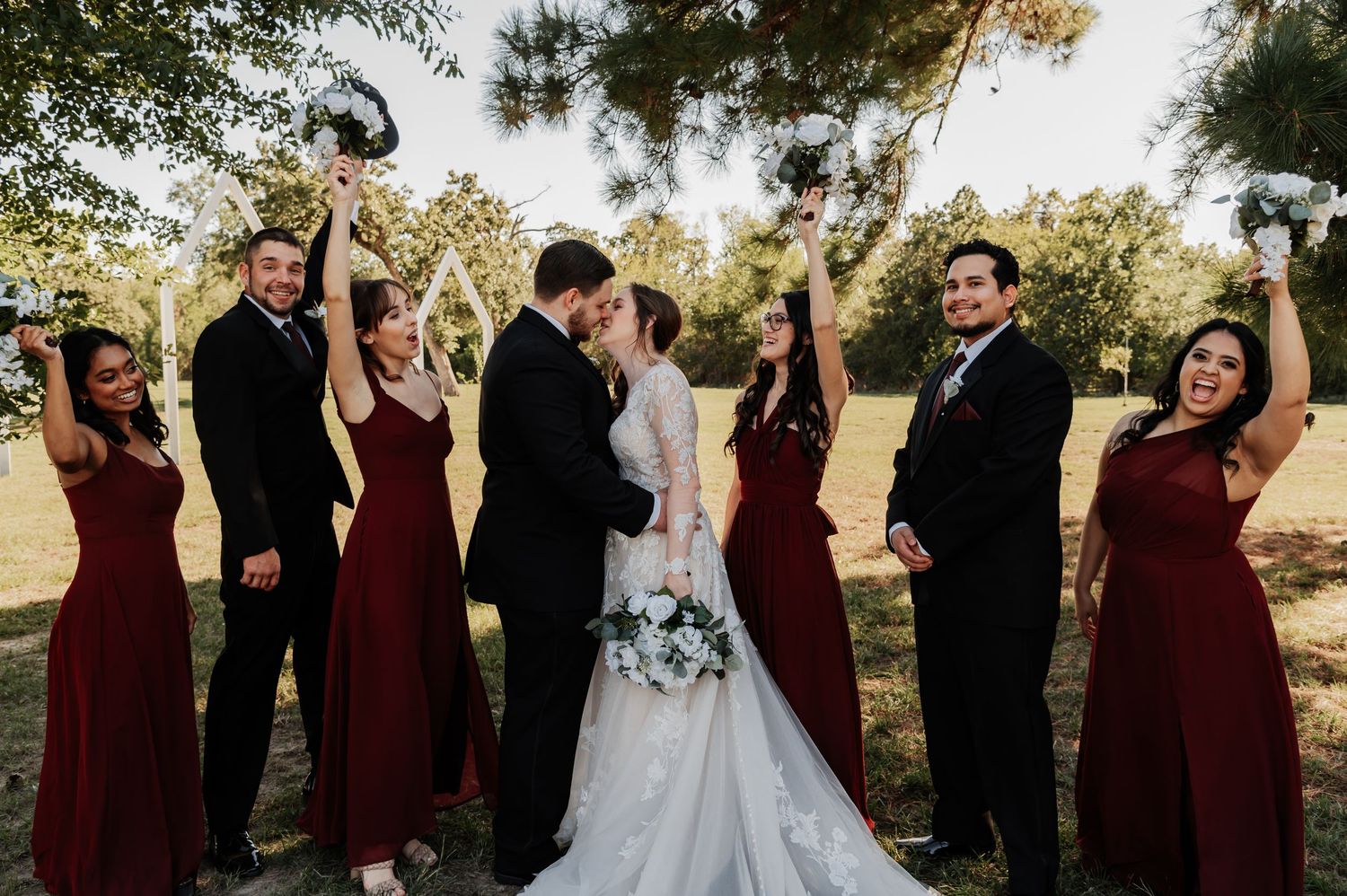 Wedding party celebrates as bride and groom kiss outdoors, bridesmaids in burgundy dresses and groomsmen in black suits cheering.