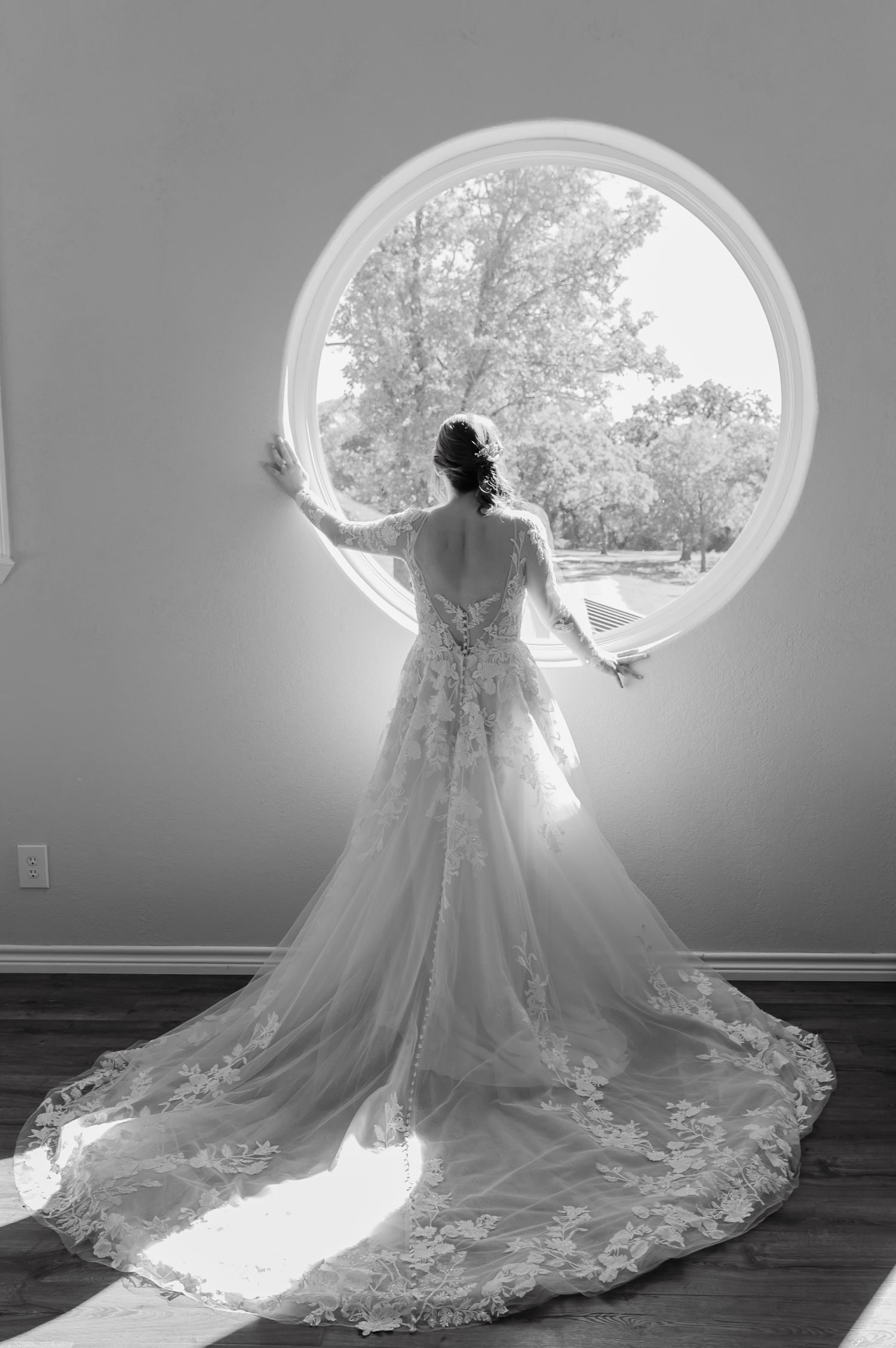 Bride in lace wedding gown with long train poses before circular window in black and white portrait.