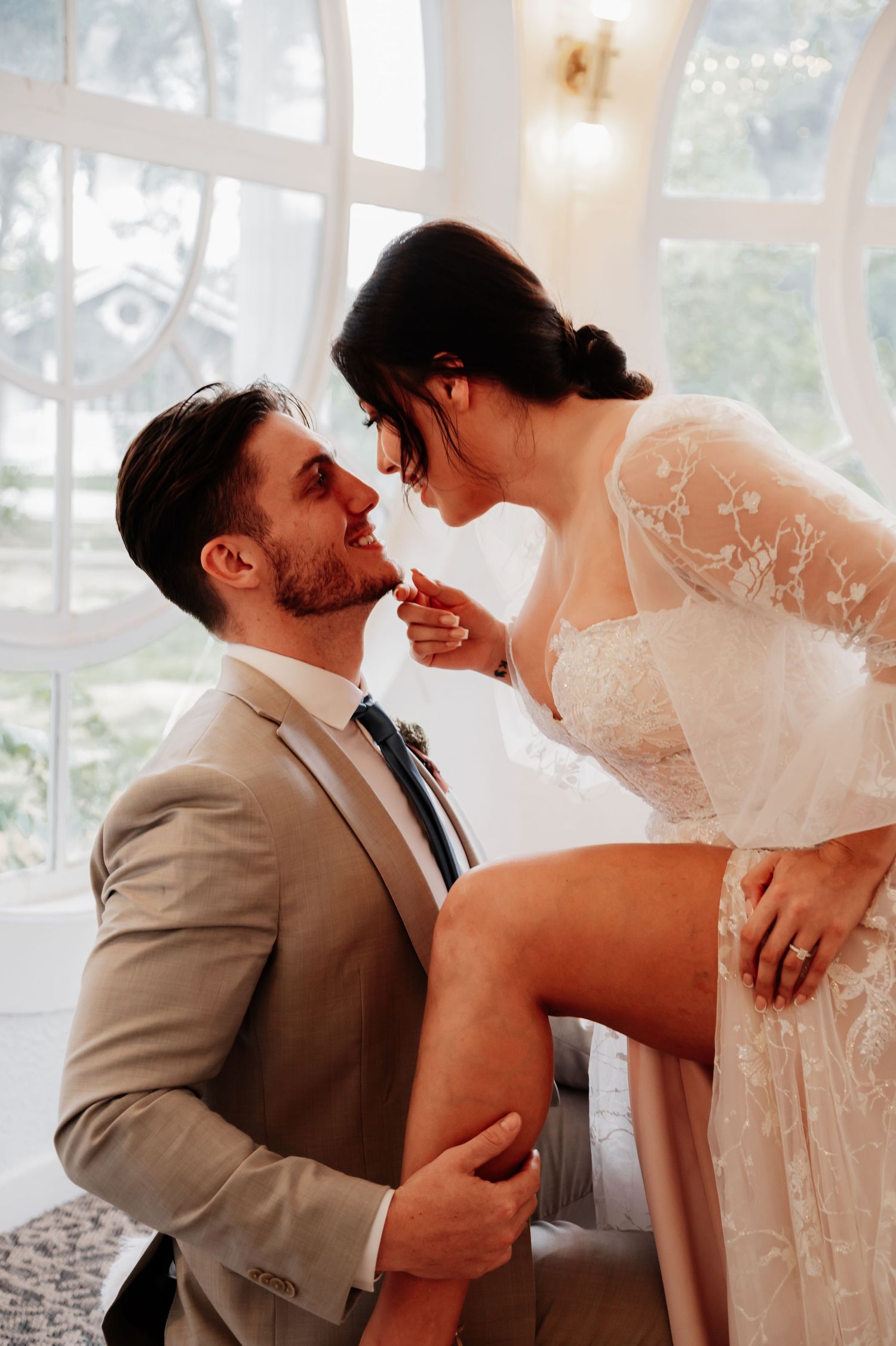 A bride and groom share a romantic moment, with the bride sitting as the groom kneels, both smiling in elegant wedding attire.