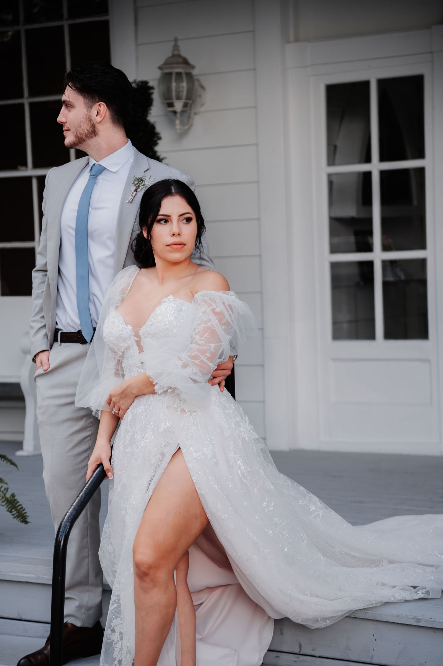 Bride in off-shoulder lace gown with slit poses on steps with groom in gray suit and blue tie at elegant white venue.