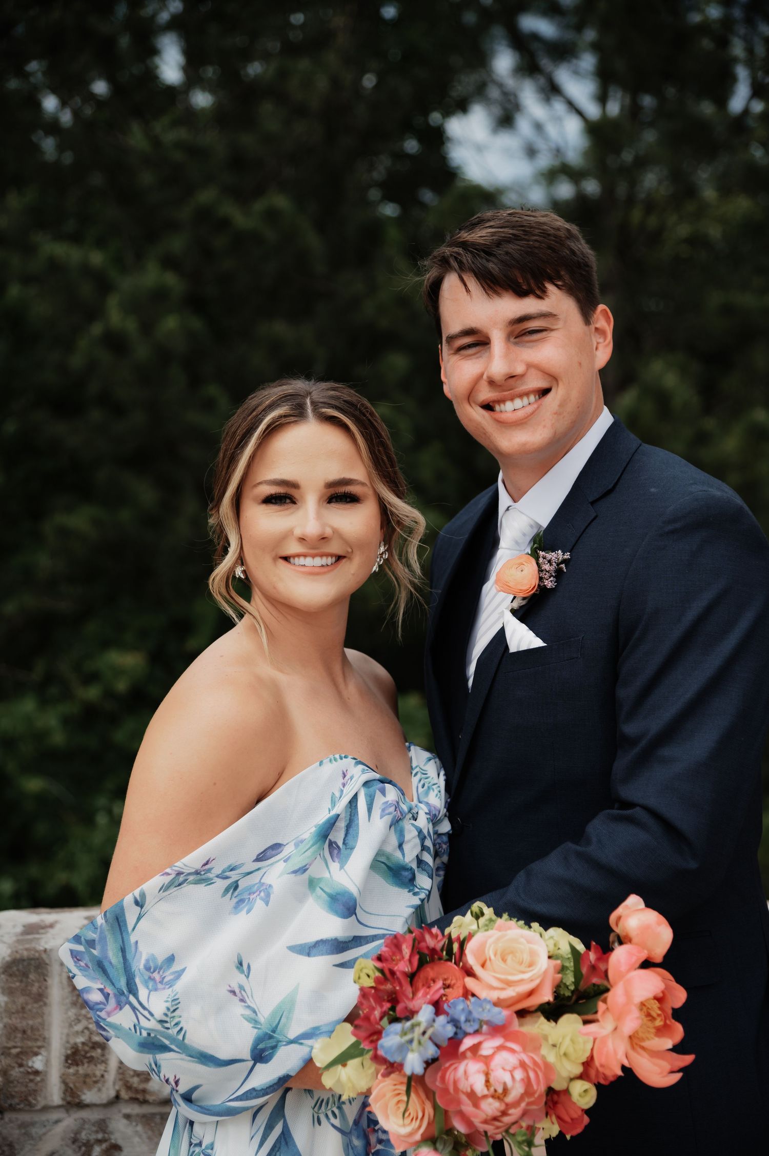 Smiling couple in wedding attire, woman in floral off-shoulder dress holding colorful bouquet, man in navy suit with boutonniere.
