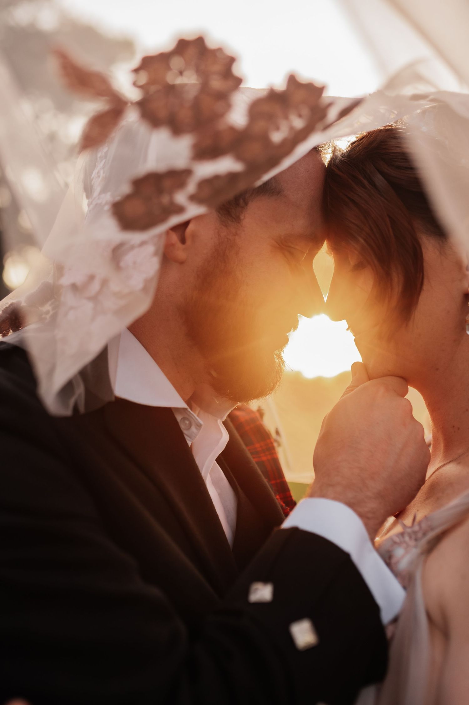 Romantic backlit wedding couple sharing an intimate moment under a floral lace veil at golden sunset.