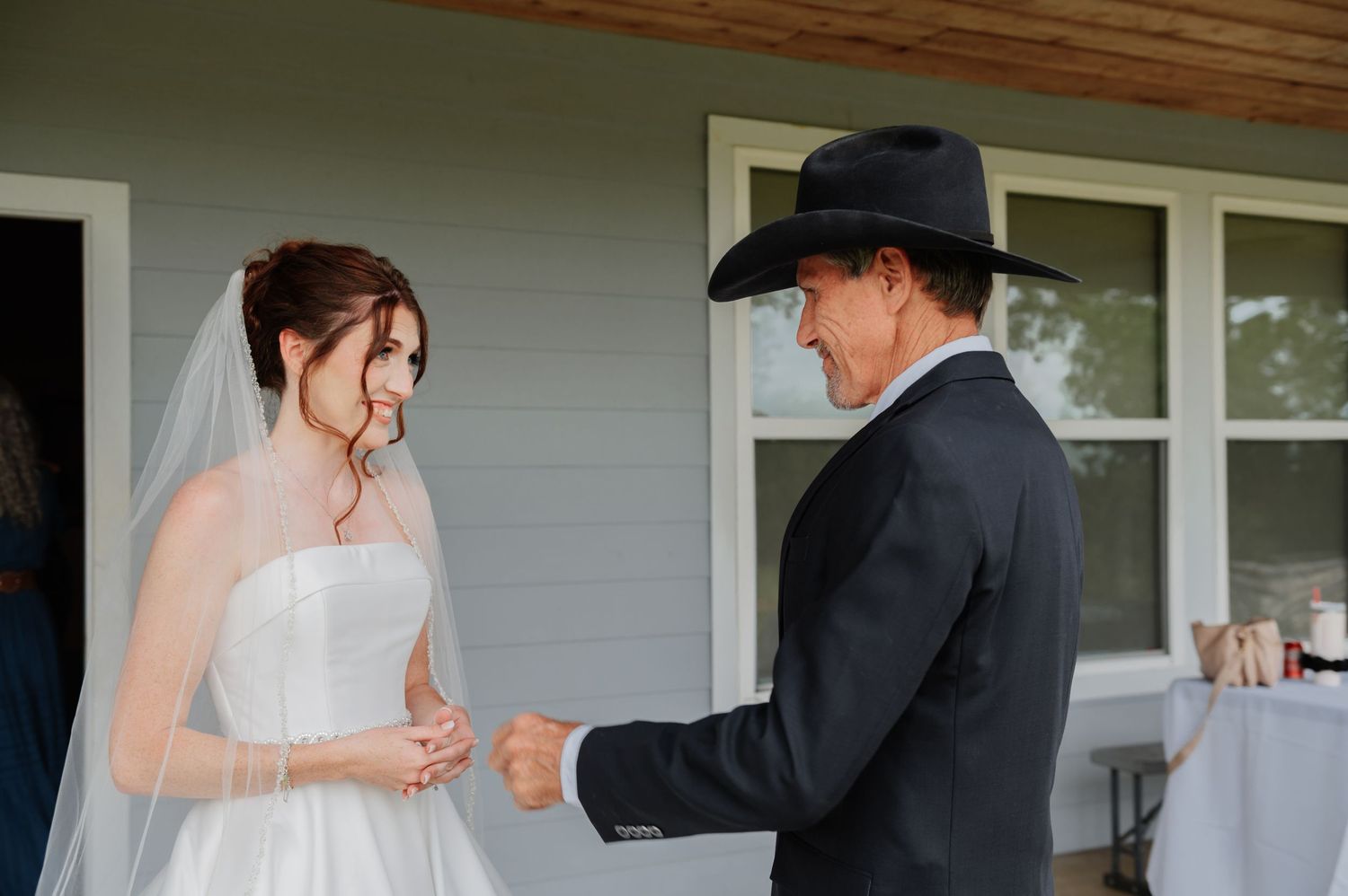 Bride in white gown and veil shares a tender first look moment with man in black suit and cowboy hat outdoors.