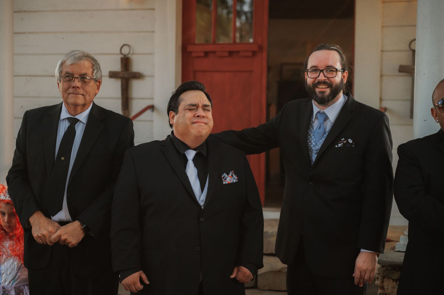 Three men in black suits and ties stand smiling outside a white chapel with a red door and cross.
