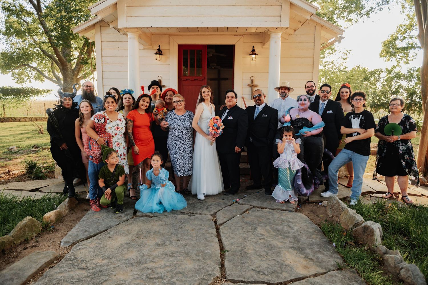 Wedding party group photo in front of a white chapel with red door at outdoor venue.