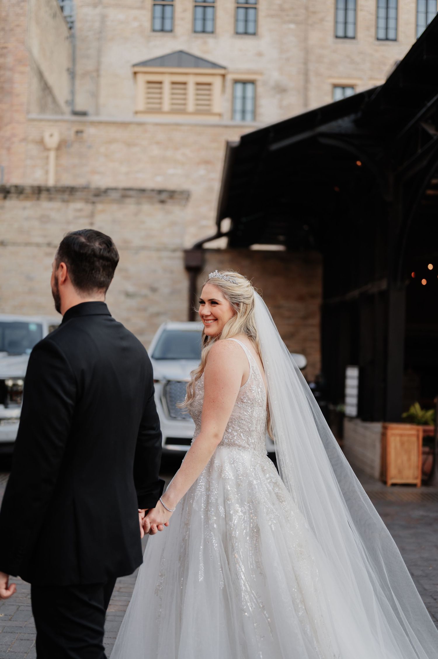 Smiling bride in beaded gown holds grooms hand walking outside historic stone building on wedding day.