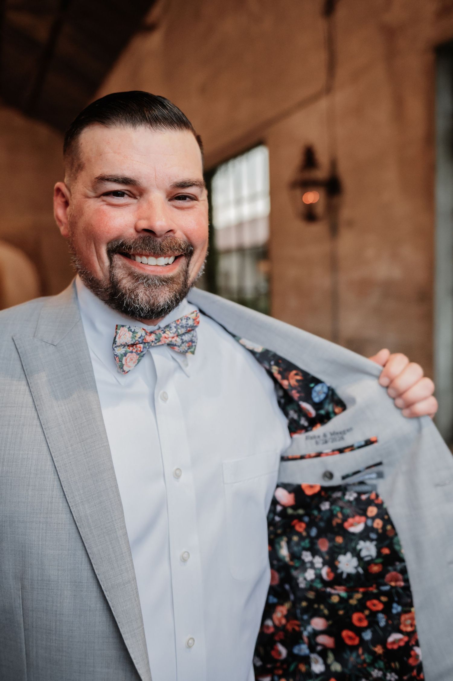 Smiling groom in light gray suit with floral bow tie showing off floral lining of jacket at wedding venue.