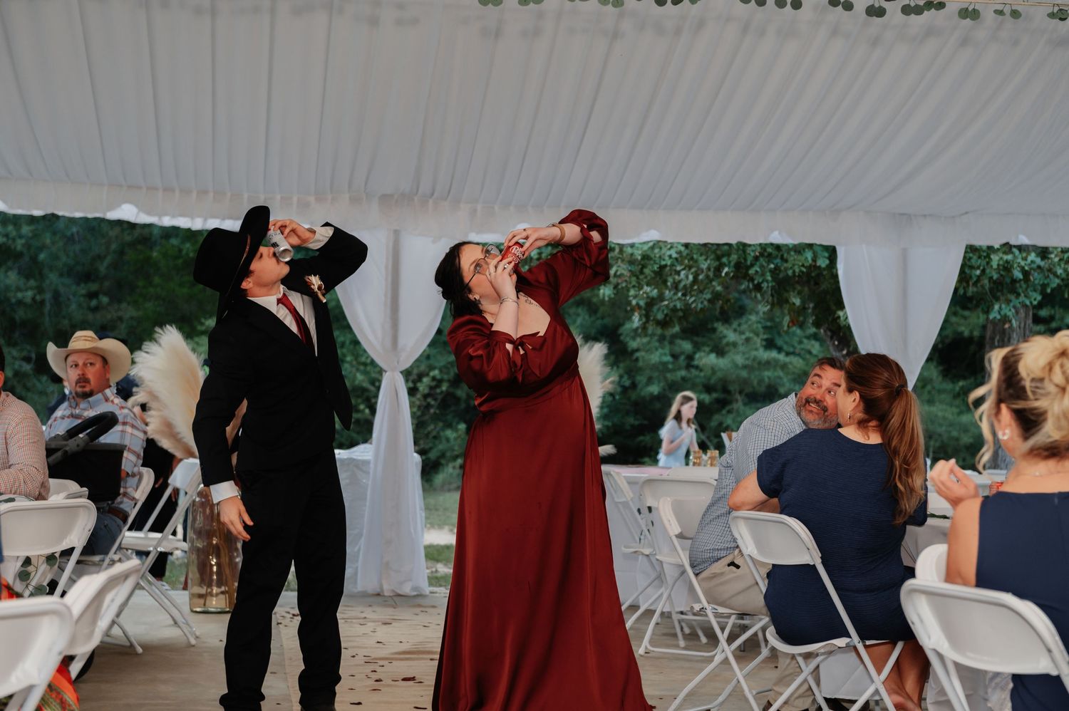 Two wedding guests in formal attire take celebratory shots during an outdoor tented wedding reception.