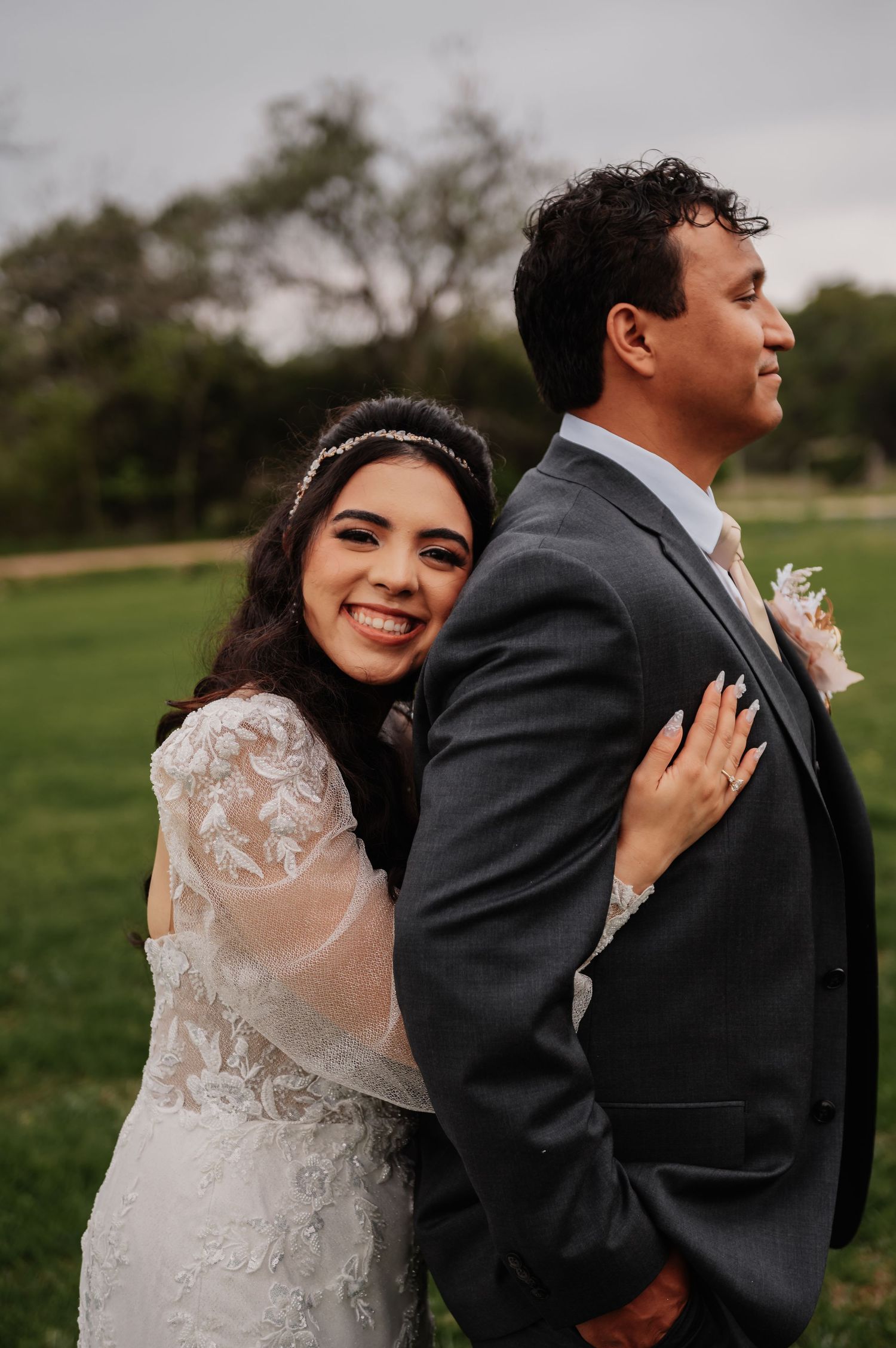 Smiling bride in lace dress embraces groom in gray suit outdoors on their wedding day.