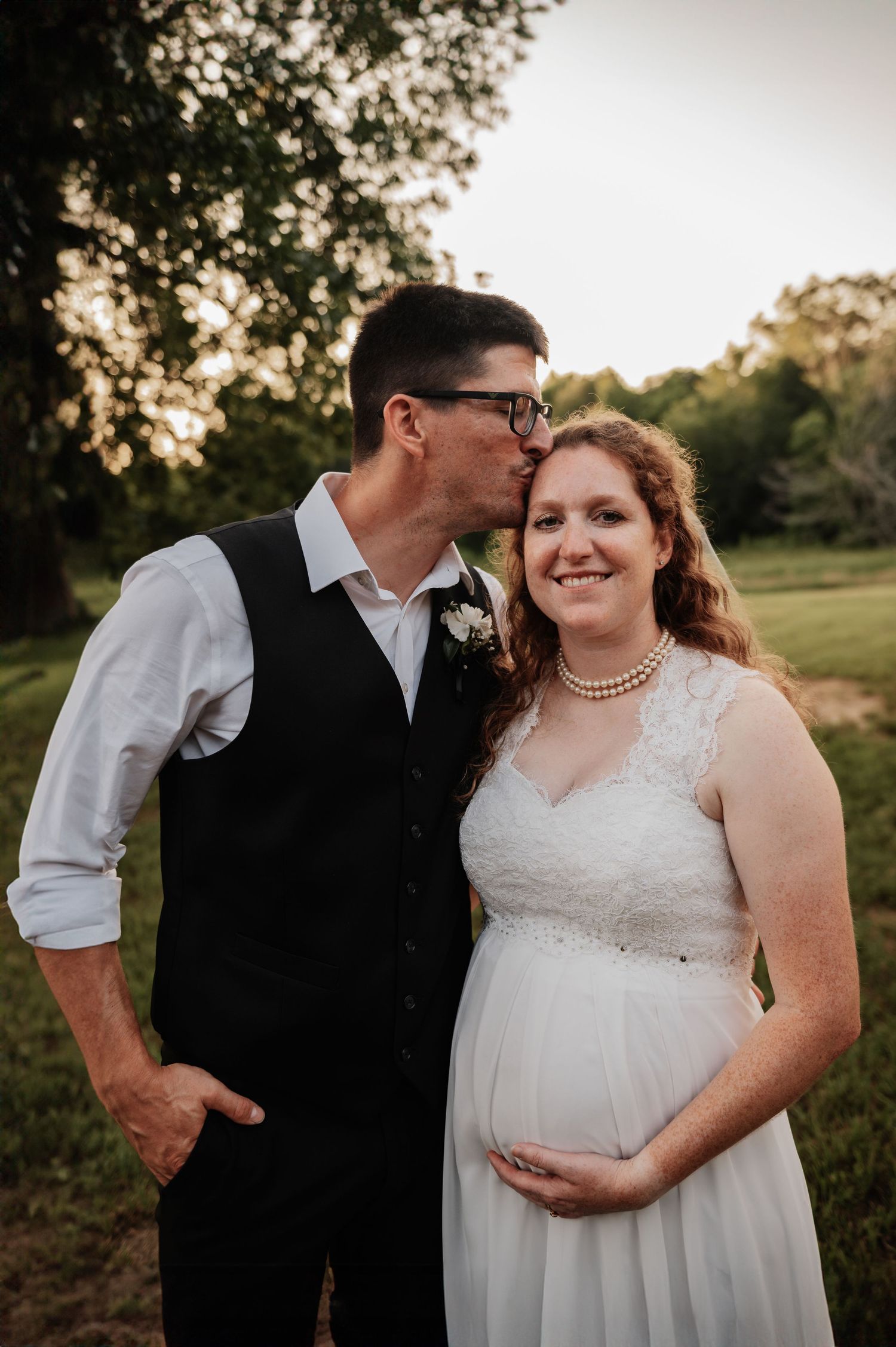 A groom kisses his pregnant bride on the cheek outdoors at golden hour, she wears a white lace gown and pearl necklace.