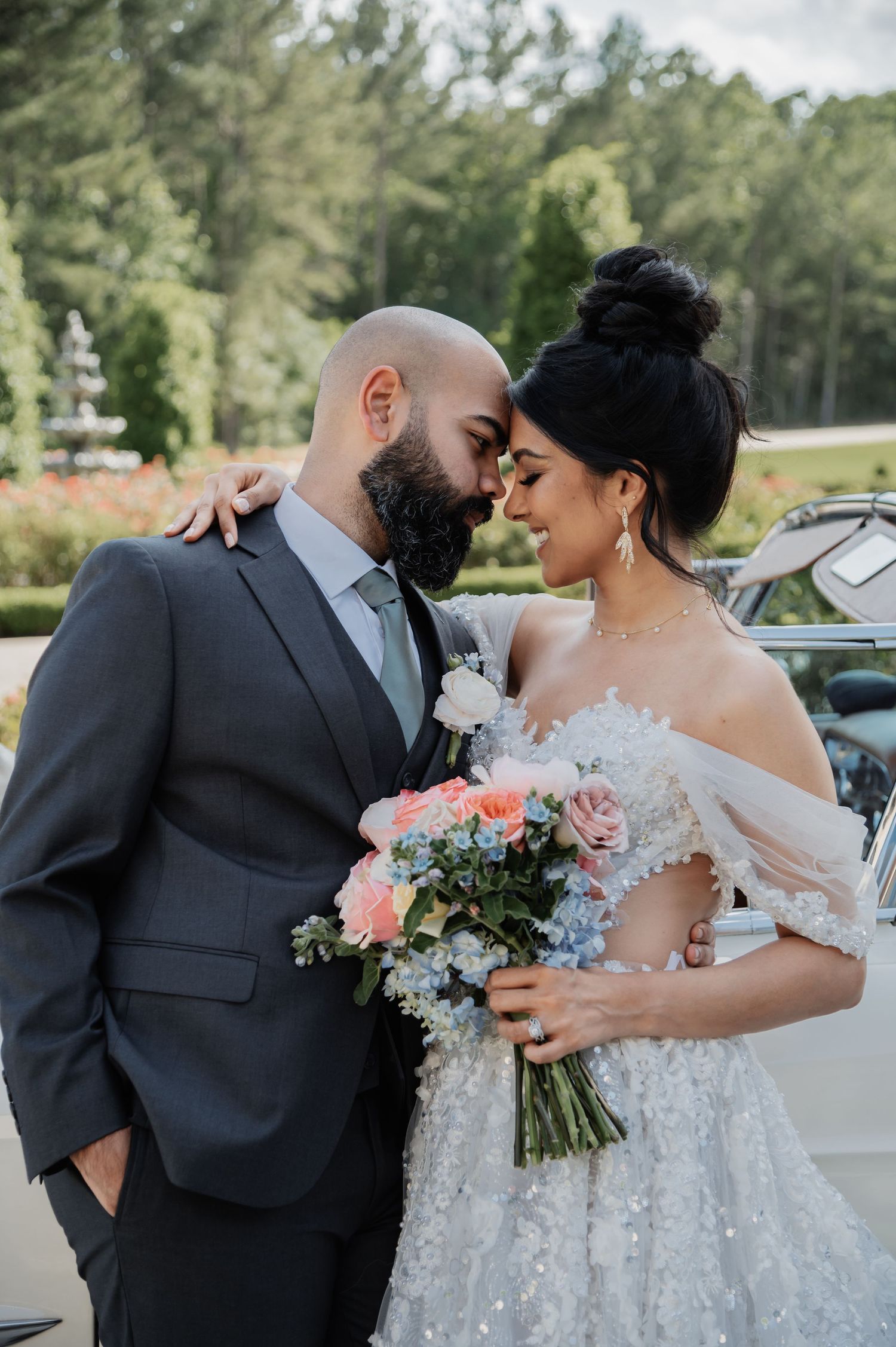 Bride and groom sharing romantic moment outdoors, bride in sparkly off-shoulder gown holding pink and blue bouquet.