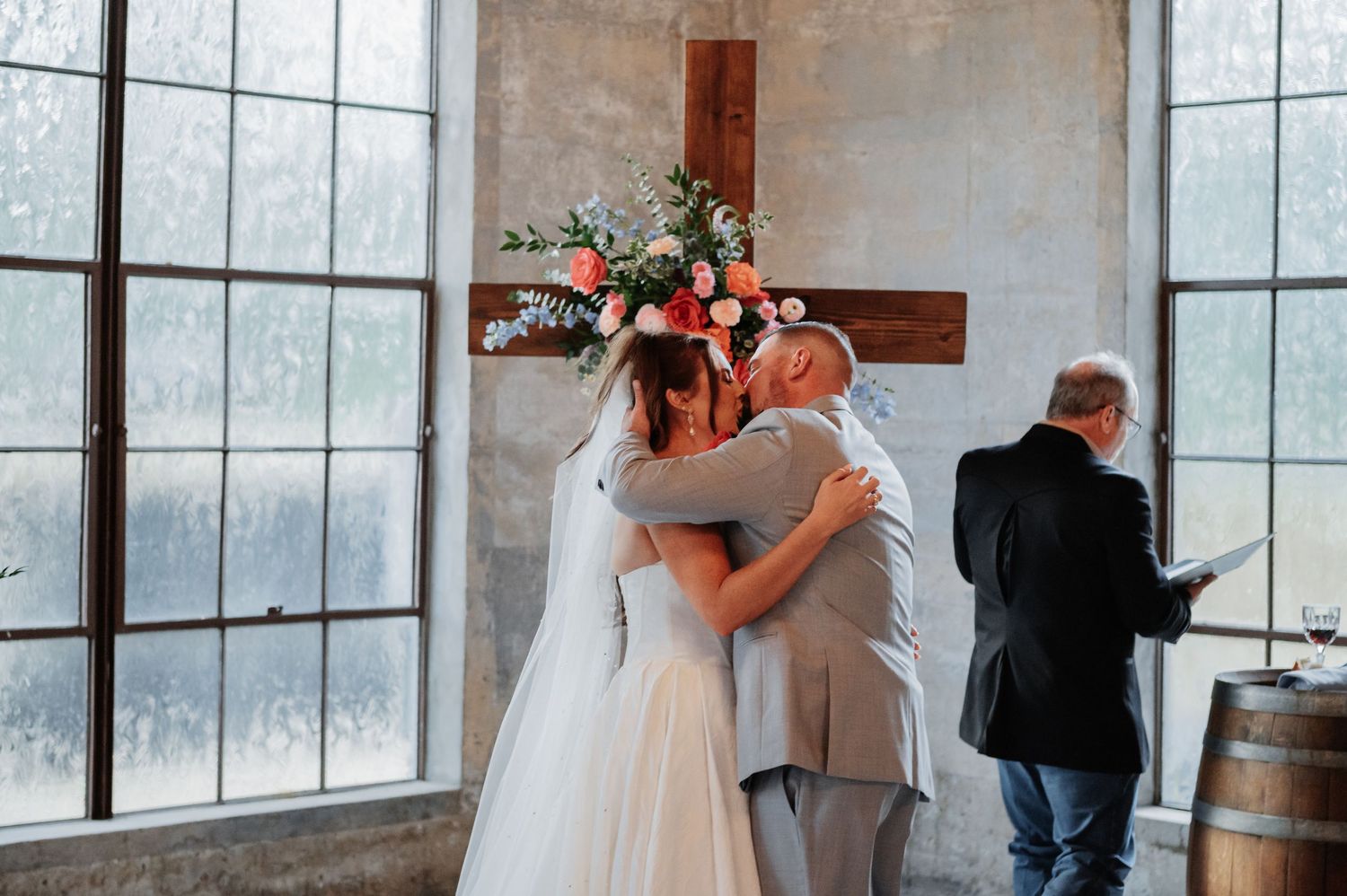 Bride and groom share an emotional hug at the altar during their wedding ceremony, with a floral cross backdrop.
