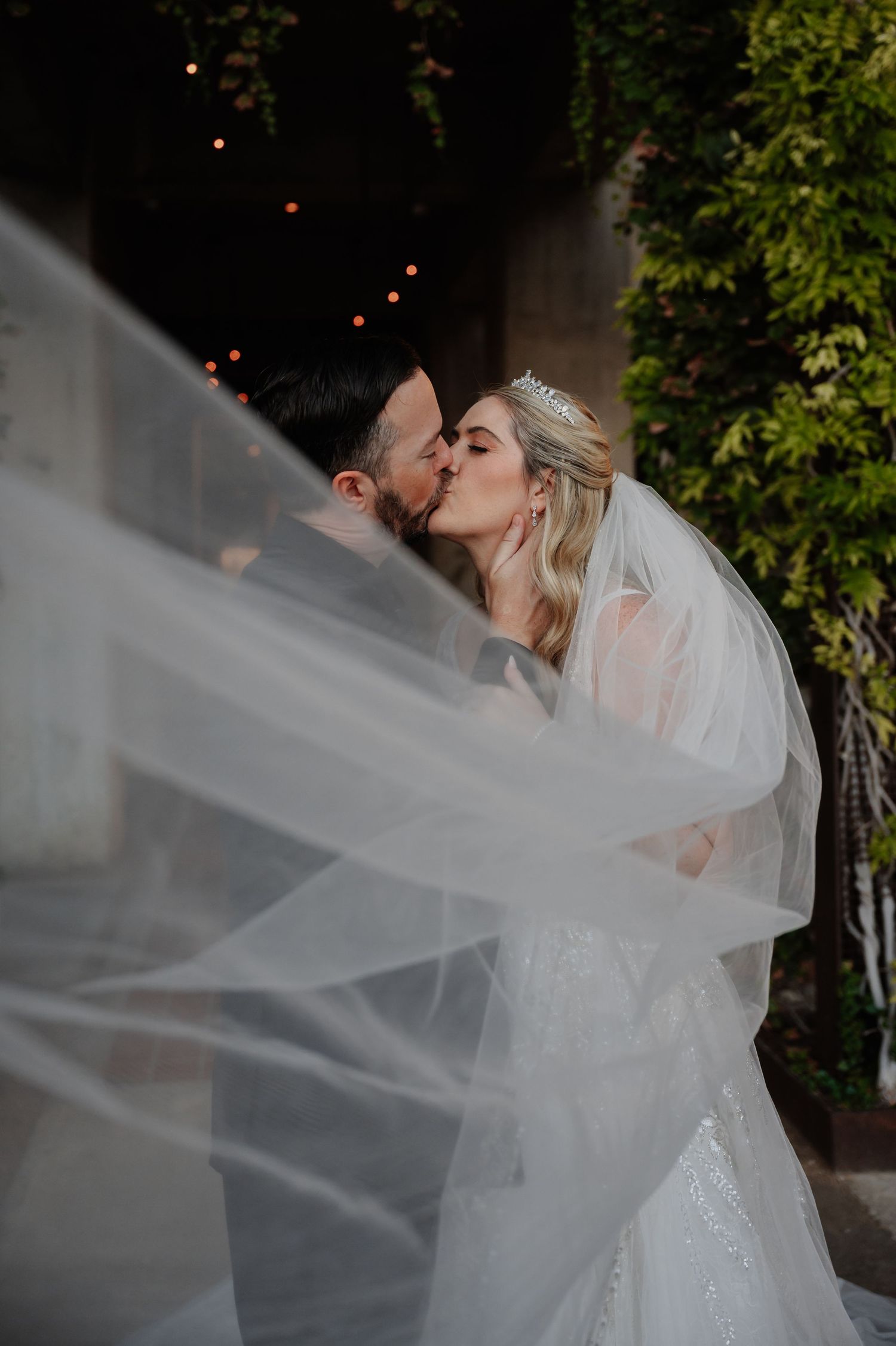 Bride and groom share a romantic kiss beneath a flowing white veil at their outdoor wedding ceremony.