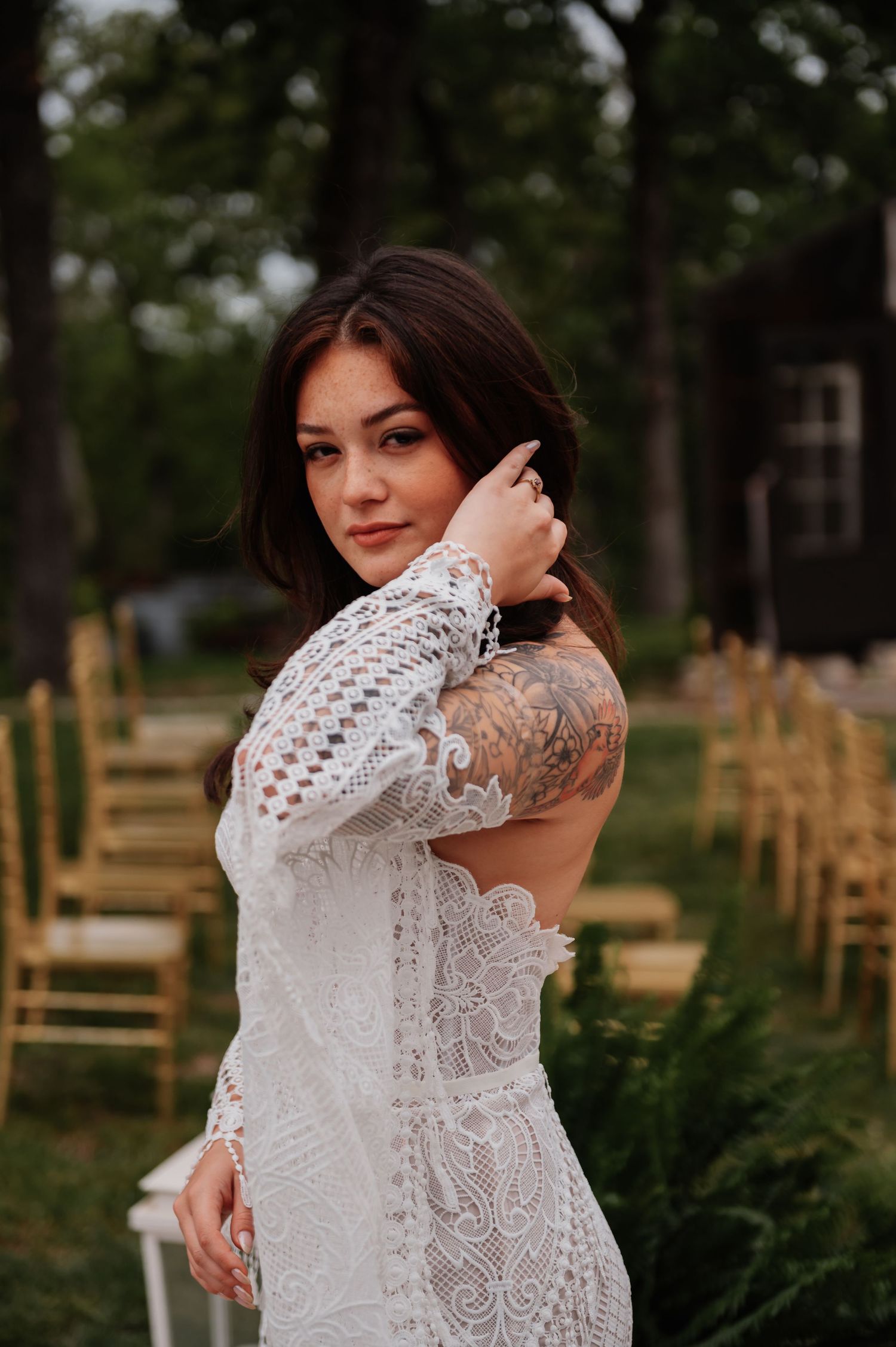 Tattooed bride in backless lace dress poses at outdoor wedding ceremony with gold chairs.