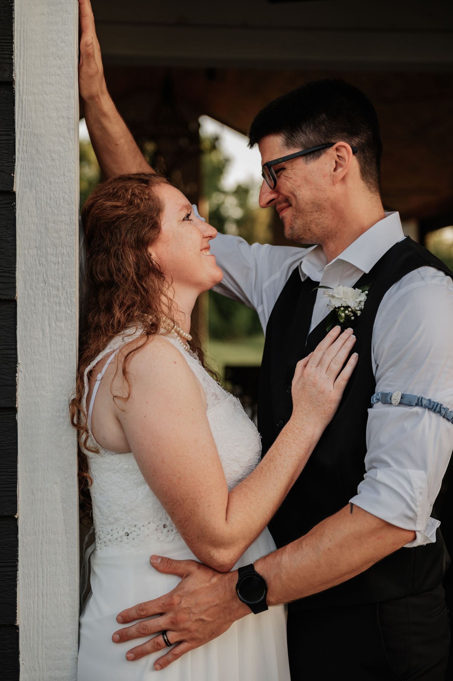 A bride and groom share a romantic moment, smiling at each other near a white pillar during their wedding day.