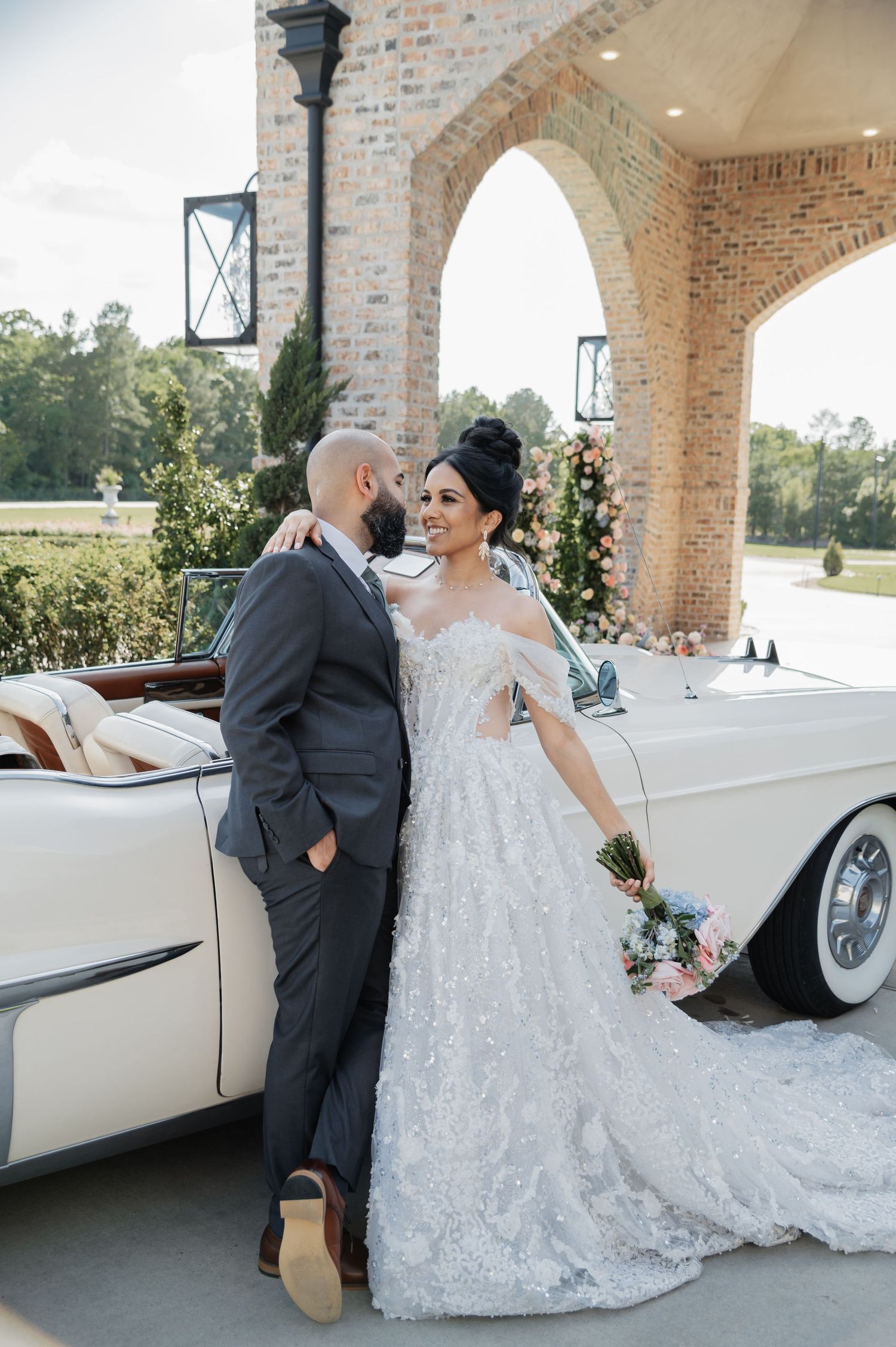 Bride in beaded gown and groom in gray suit pose by vintage white convertible at elegant brick arch venue.