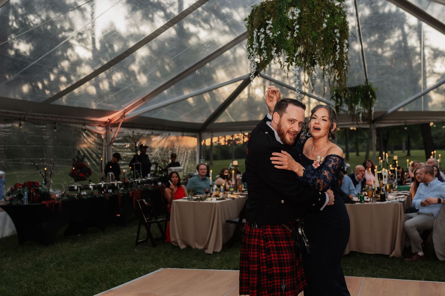 Couple shares first dance under clear tent at outdoor wedding reception, groom in red tartan kilt.