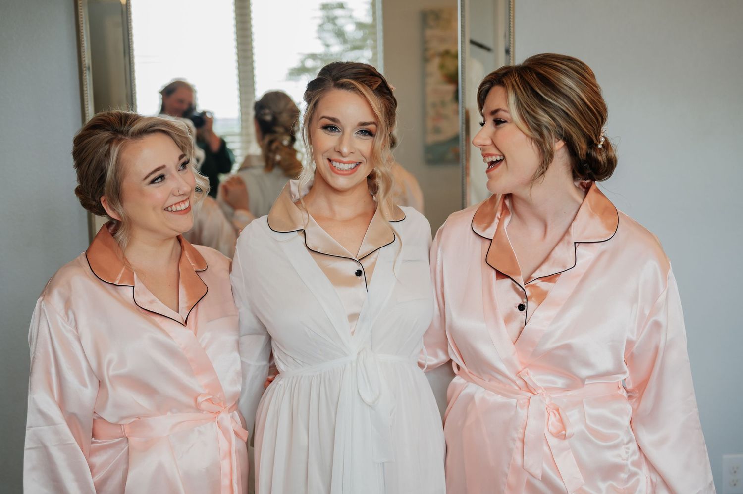 Bride and two bridesmaids in matching pink satin robes smiling together during wedding morning preparations.