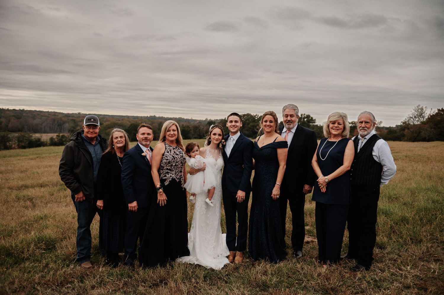 Wedding party poses in a rustic field, bride in white gown surrounded by family in dark formal attire under overcast skies.