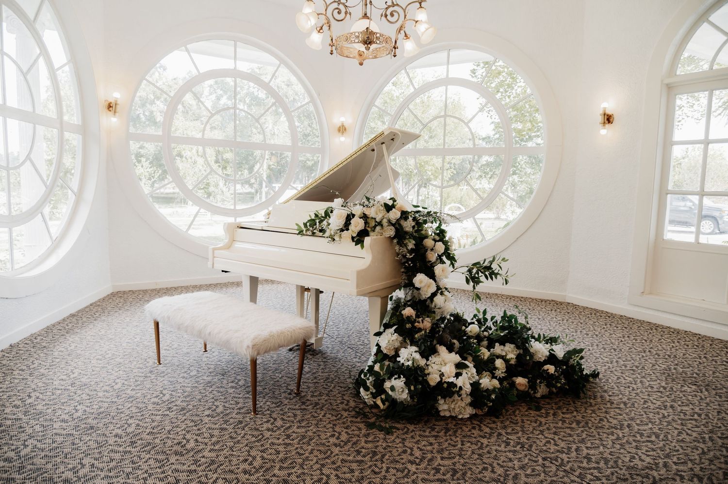 White grand piano adorned with lush floral arrangements in an elegant white room with arched windows.