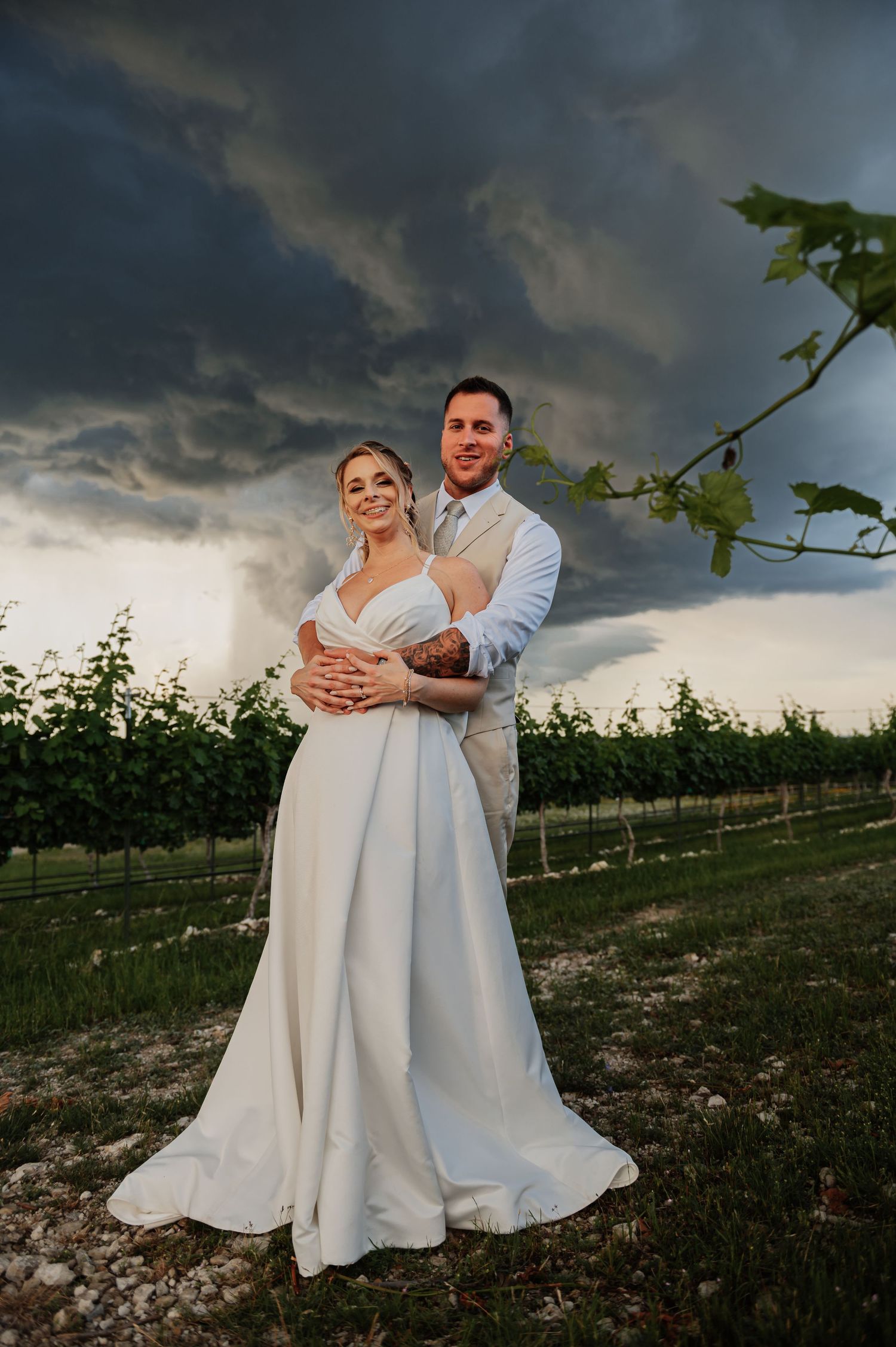 Bride and groom embrace in a vineyard under dramatic storm clouds during their outdoor wedding ceremony.
