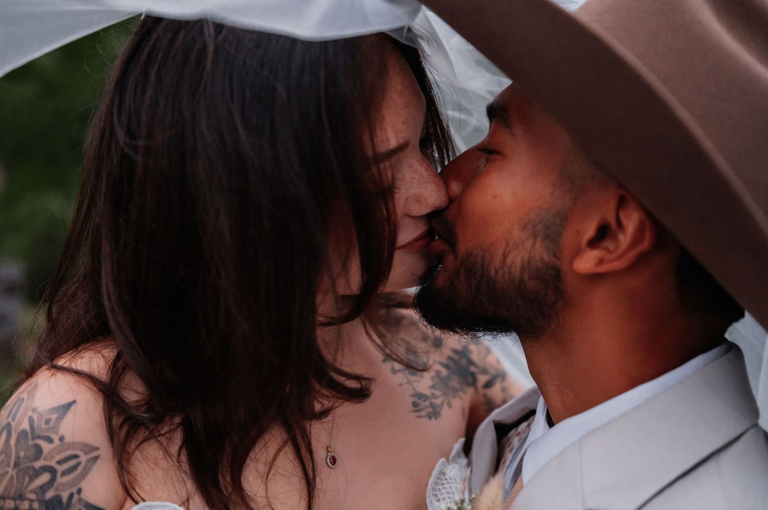Tattooed couple sharing a romantic kiss under a veil at their wedding ceremony.