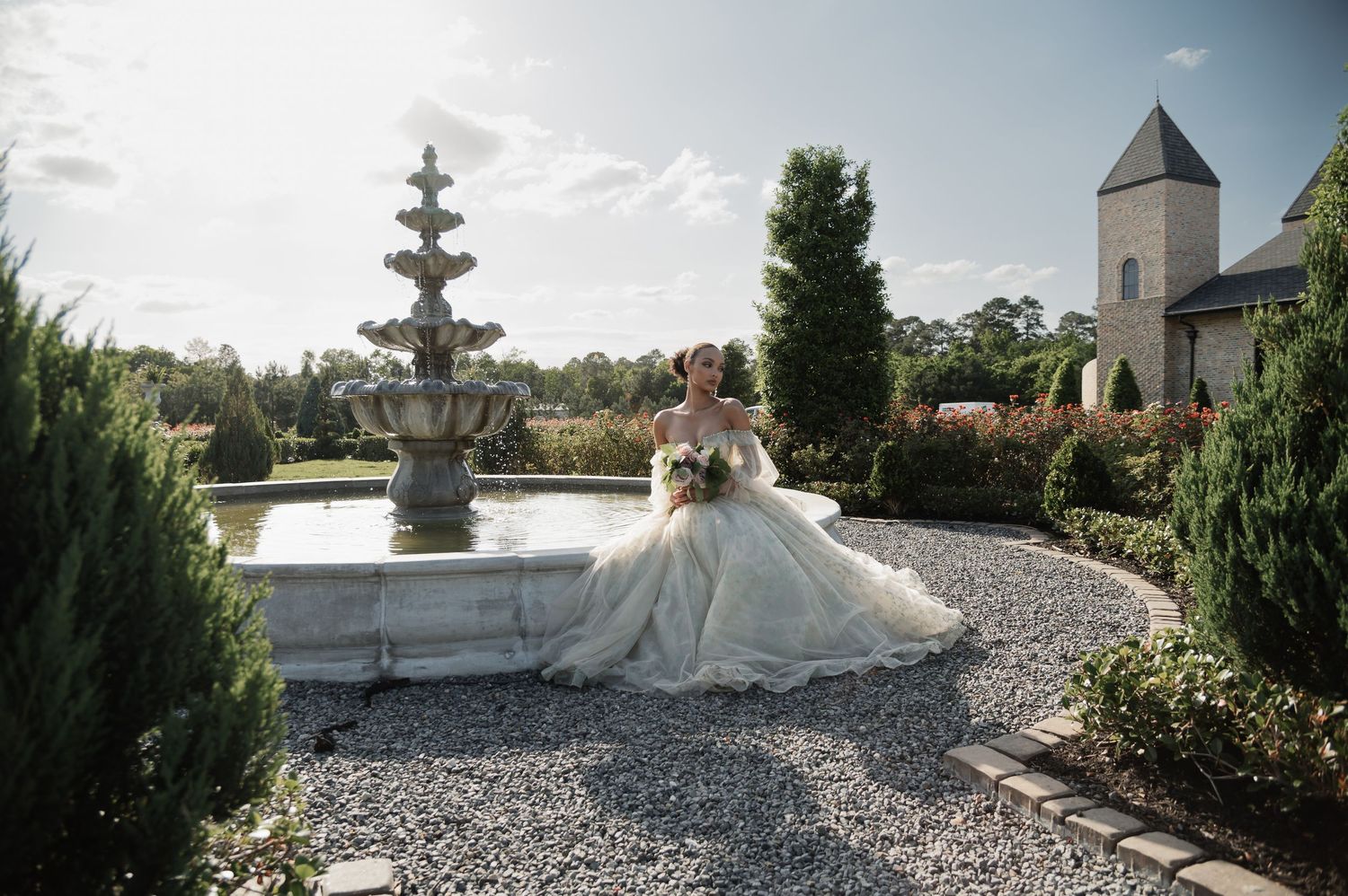 Bride in a flowing white ball gown poses by a stone fountain in an elegant garden with lush greenery.