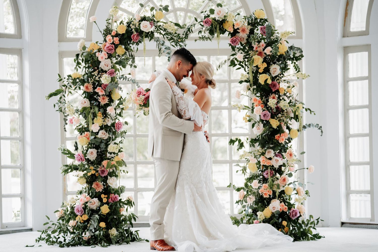 Bride and groom embrace under a stunning floral arch with pink, white, and peach blooms in an elegant bright venue.