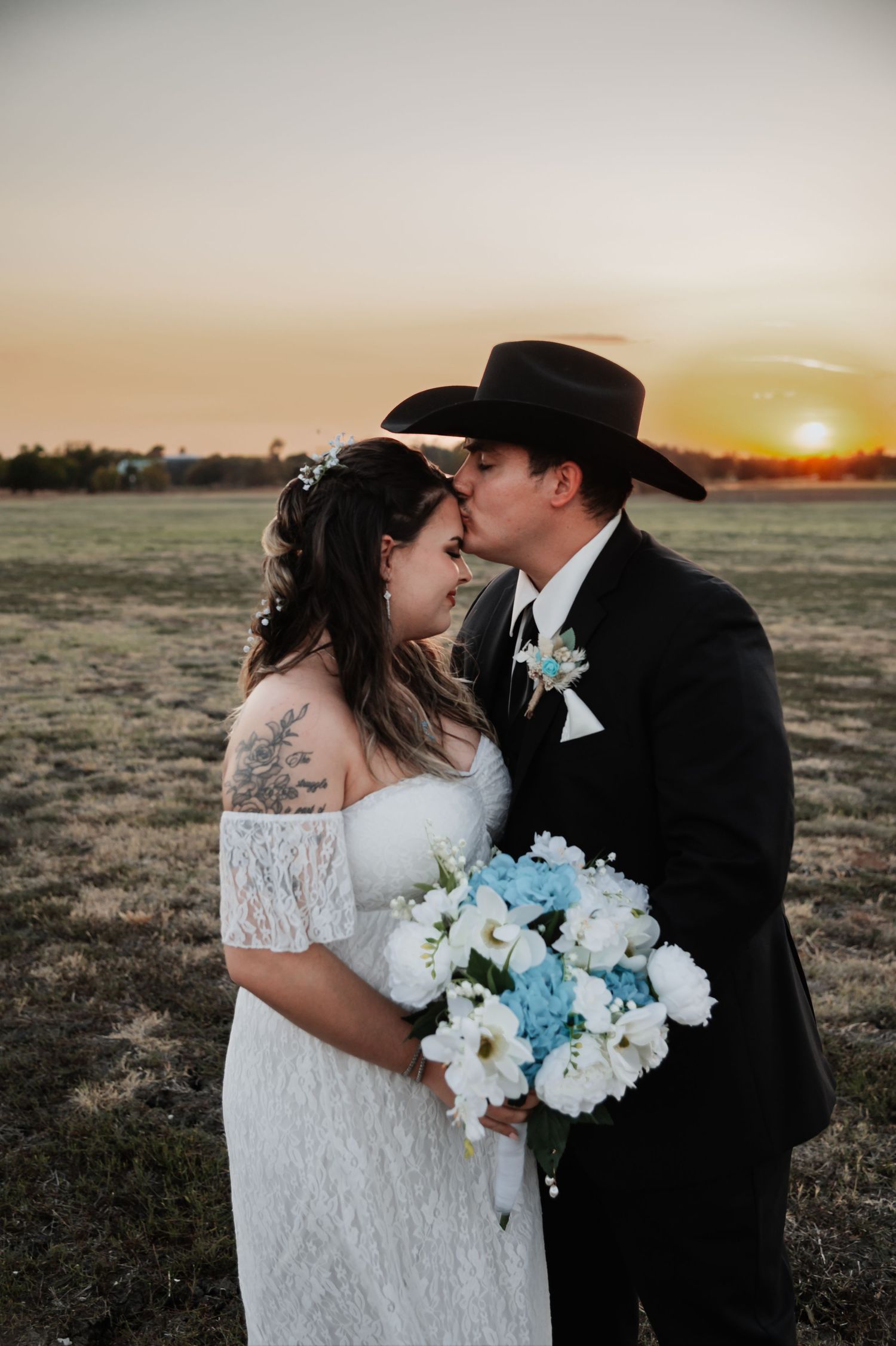 Newlywed couple sharing a kiss at sunset, bride in lace gown holding blue bouquet, groom in black suit and cowboy hat.