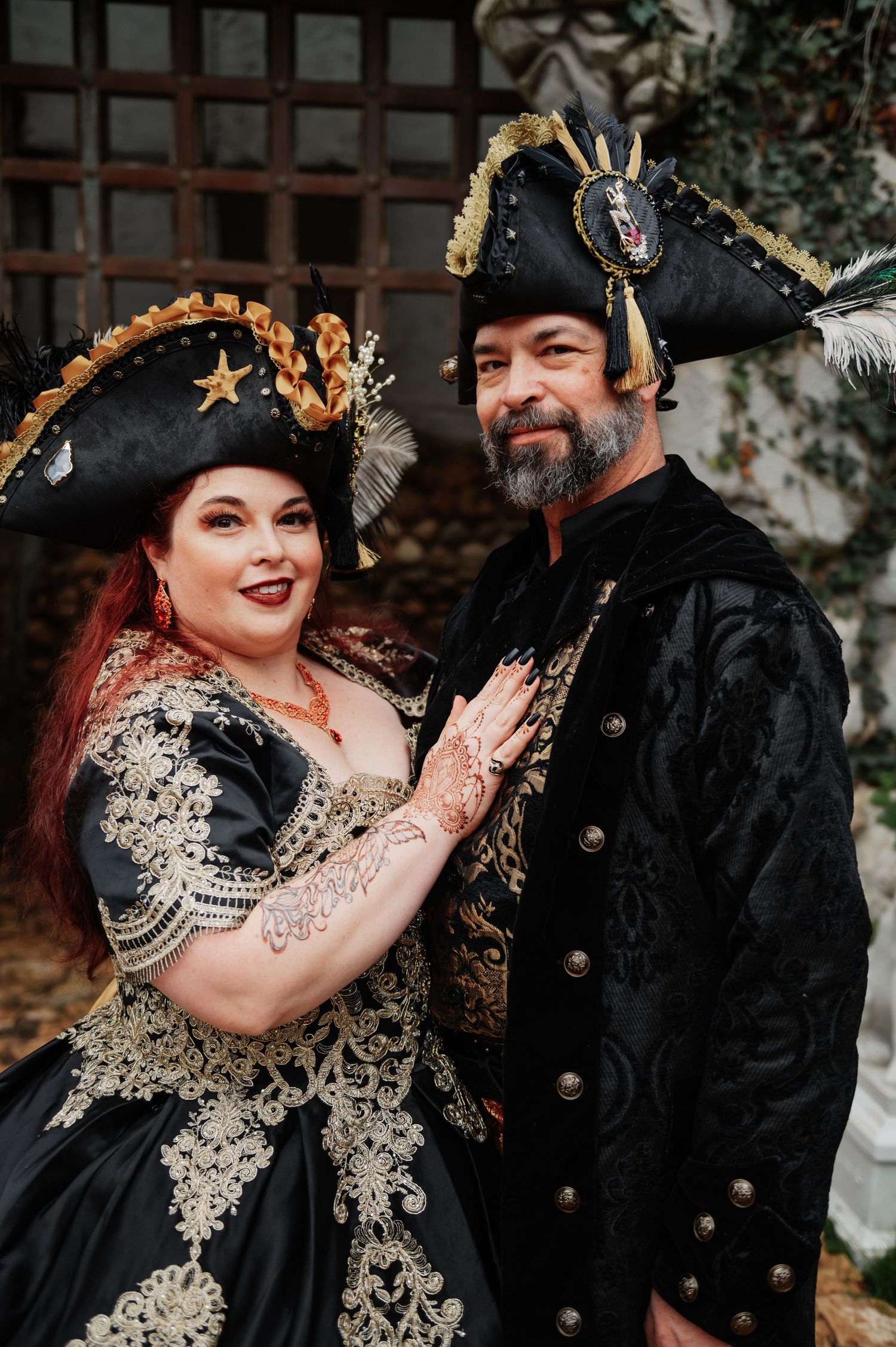 Couple in elegant black and gold pirate costumes with tricorn hats posing together at a themed event.