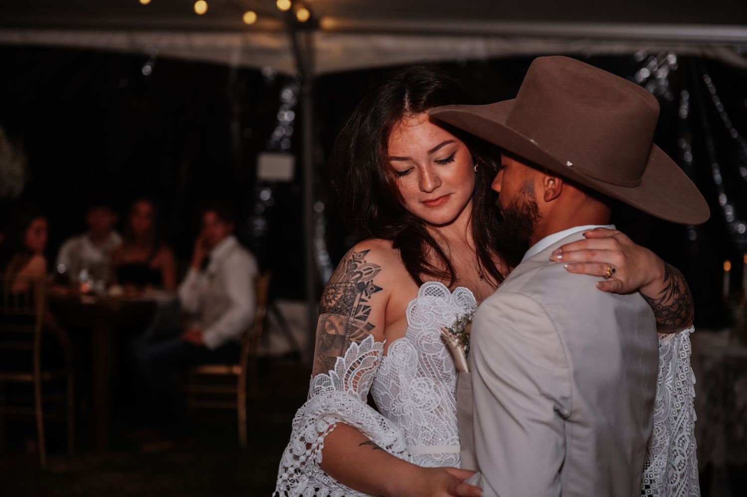 Bride and groom share first dance under tent lights, she wears lace gown with tattoos, he wears brown cowboy hat.