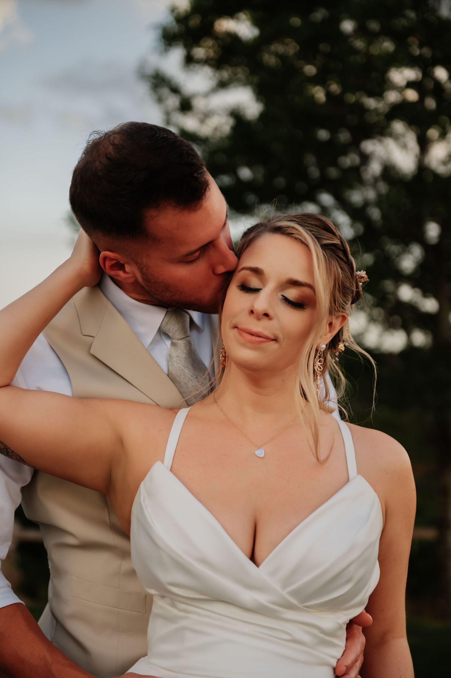 Groom in tan vest kissing bride on cheek as she closes her eyes in romantic outdoor wedding portrait.