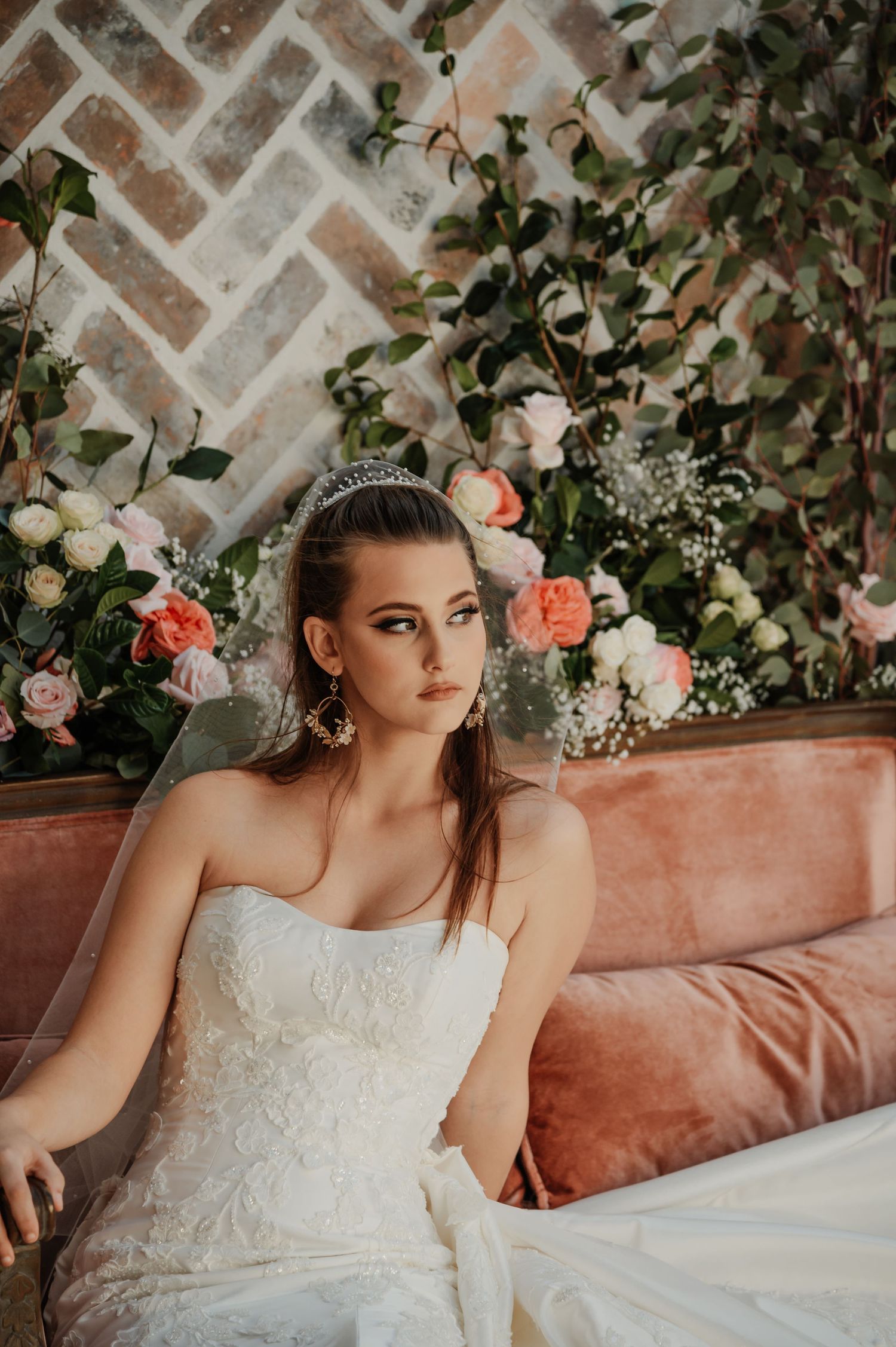 Beautiful bride in strapless lace gown wearing tiara sits on pink velvet sofa surrounded by floral arrangements.