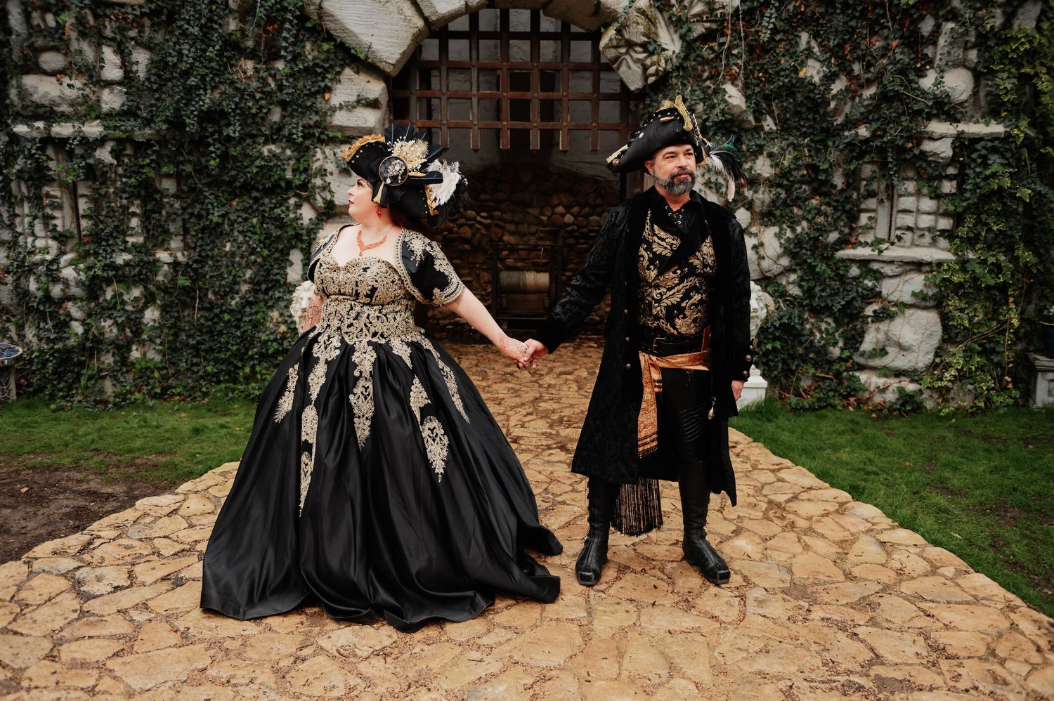 Bride and groom in black gold medieval wedding costumes holding hands outside stone castle wall.