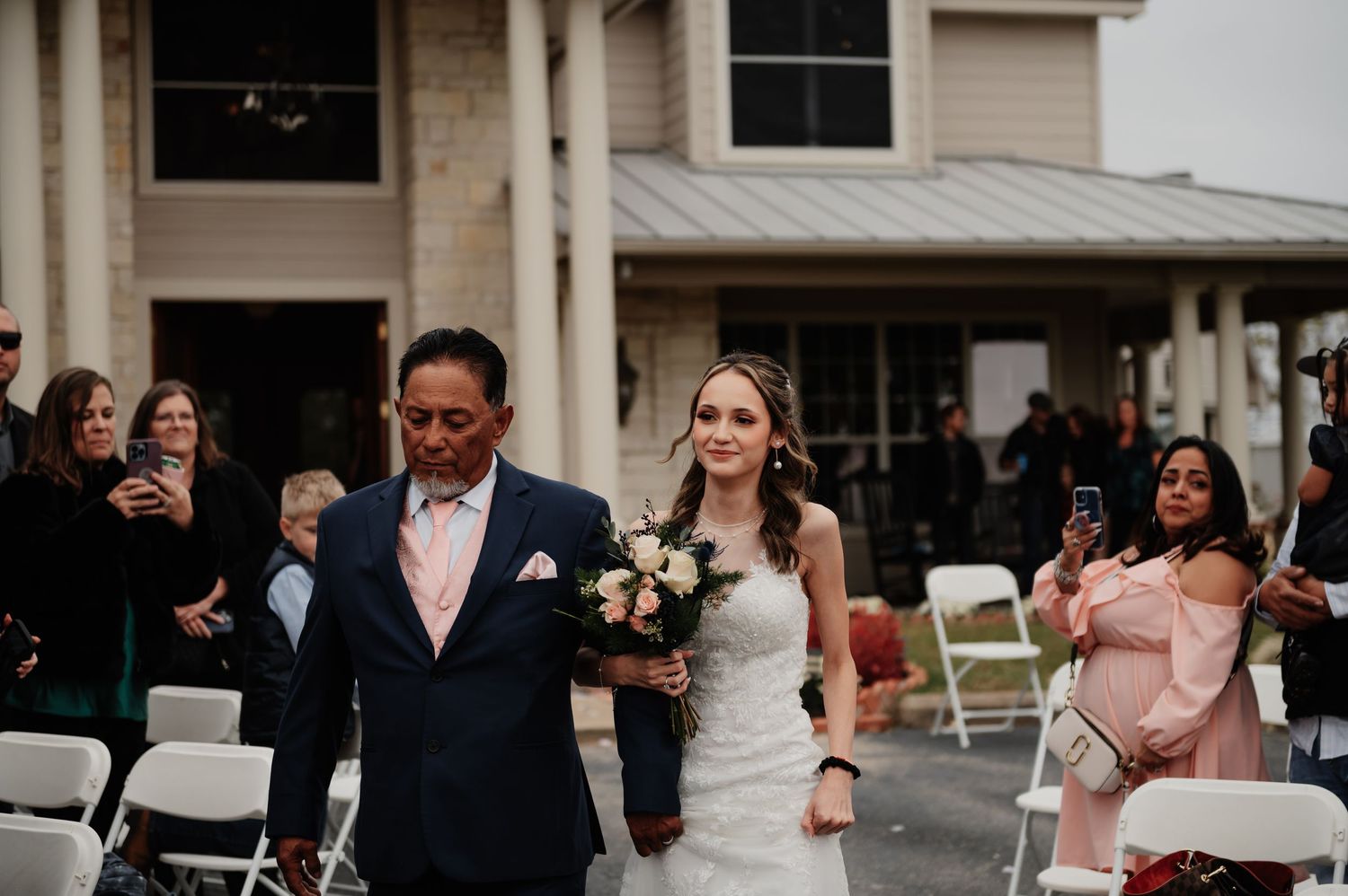 Bride walking down the aisle with her father at an outdoor wedding ceremony, holding a bouquet.