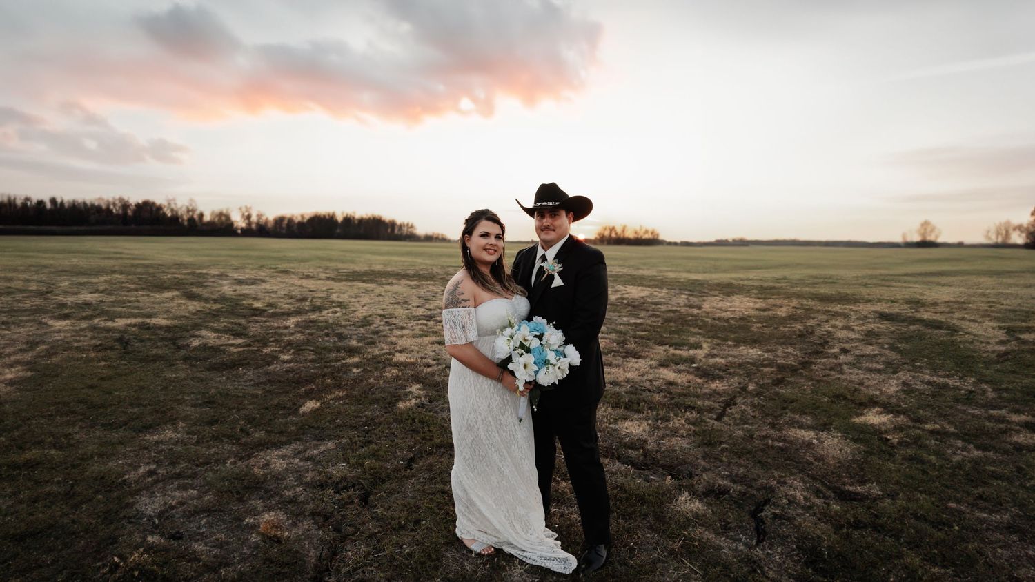 Bride and groom posing in open field at sunset, groom wearing black cowboy hat, bride holding blue bouquet.