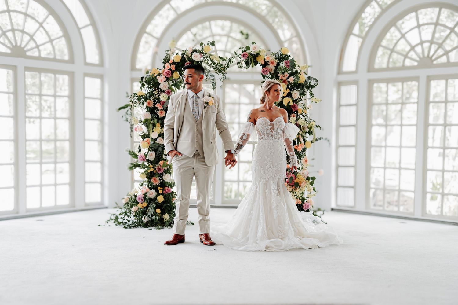 Bride and groom holding hands before a colorful floral arch in an elegant white venue.