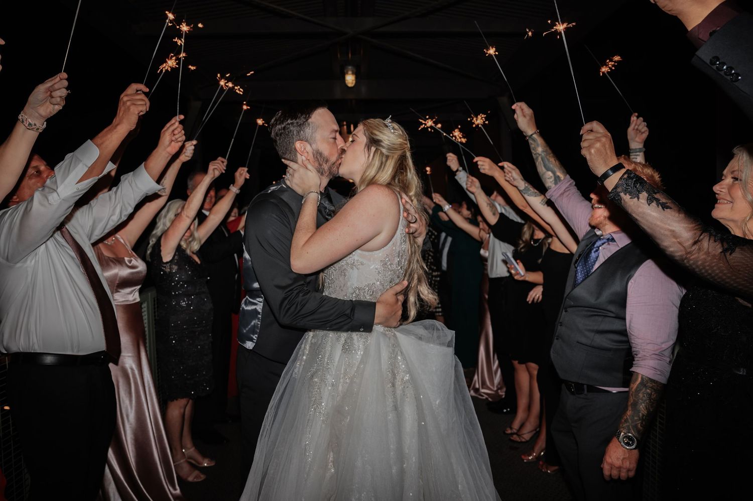 Bride and groom share a romantic kiss during a sparkler send-off surrounded by wedding guests at their reception.